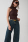 A woman modeling a sleeveless dark brown mock-neck top paired with dark blue wide-leg jeans and a gold belt, set against a plain light gray studio background.