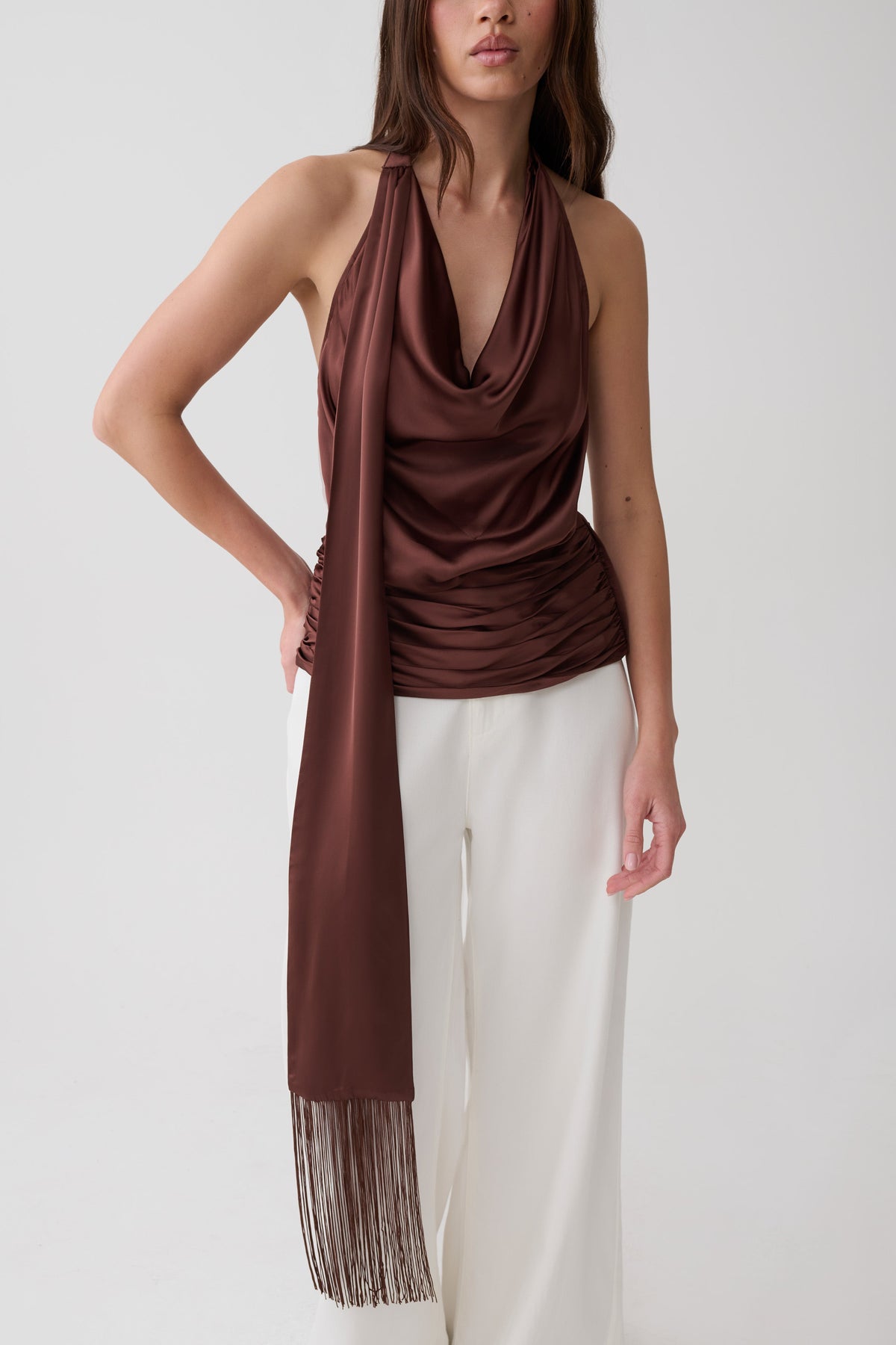 A woman modeling a dark brown draped halter top with a matching long scarf draped over her shoulder, paired with white wide-leg pants, against a plain light gray studio background.