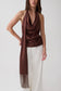 A woman modeling a dark brown draped halter top with a matching long scarf draped over her shoulder, paired with white wide-leg pants, against a plain light gray studio background.